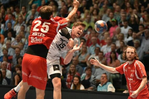 Zwei männliche Handballspieler in Aktion; der eine in einem roten Trikot blockt einen Gegner in einem weißen Trikot, der den Ball hält, während ein anderer Spieler in Rot zusieht. Der Hintergrund zeigt eine überfüllte Arena mit Zuschauern.