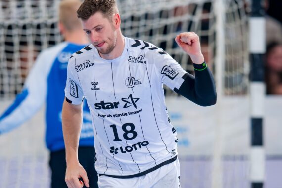 Ein männlicher Handballspieler in einem weißen Trikot mit der Nummer 18 steht während eines Spiels vor einem Tor und feiert mit geballter Faust.