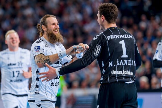 Zwei männliche Handballspieler, einer in einem weißen und einer in einem schwarzen Trikot, schütteln sich auf dem Spielfeld lächelnd die Hände, während im Hintergrund eine jubelnde Menge steht.