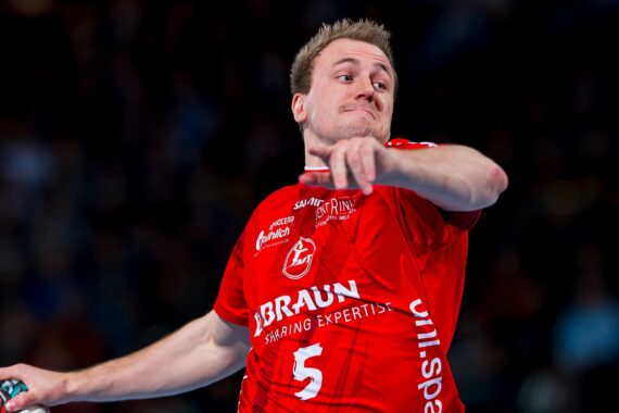 Ein männlicher Handballspieler im roten Trikot mit der Nummer 5 bereitet sich darauf vor, den Ball während eines Spiels zu werfen, mit einem konzentrierten Gesichtsausdruck und einer unscharfen Menschenmenge im Hintergrund.