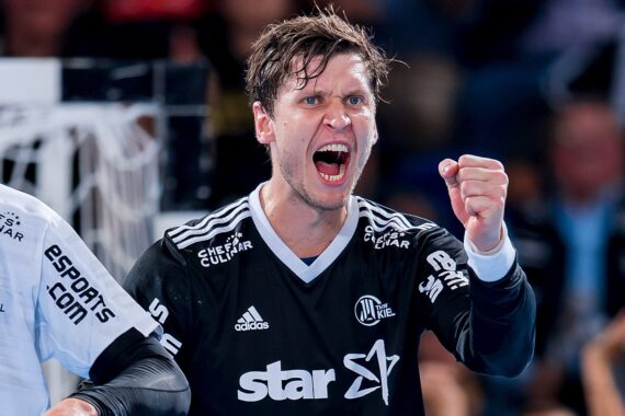 Ein männlicher Handballspieler in einem schwarzen Trikot ballt seine Faust und schreit leidenschaftlich, während er ein Spiel feiert. Im Hintergrund sind unscharfe Mannschaftskameraden und Zuschauer zu sehen.