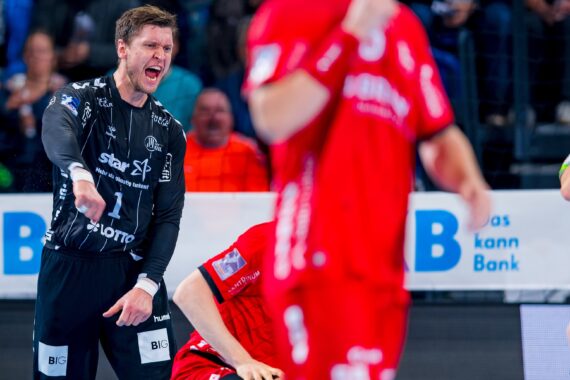 Ein Handballspieler in einem schwarzen Trikot schreit und gestikuliert leidenschaftlich, während Spieler in roten Trikots während eines Spiels in der Nähe des Spielfelds stehen. Im unscharfen Hintergrund sind Zuschauer zu sehen.
