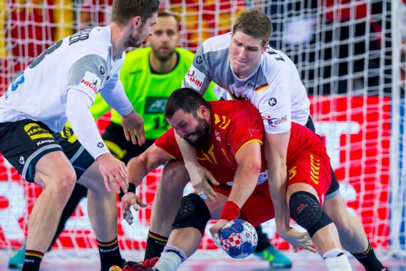 Ein Handballspieler in einem roten Trikot kämpft darum, den Ball unter Kontrolle zu halten, während er von zwei Spielern in weißen Trikots eng verteidigt wird.