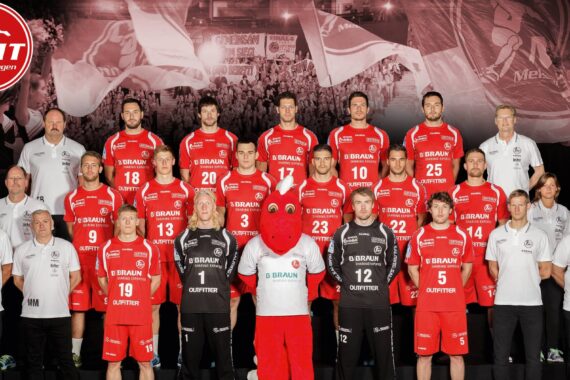Eine Männerhandballmannschaft in roten Trikots mit Trainern in weißen Hemden steht und sitzt in Reihen. Ein rotes Maskottchen steht in der vorderen Mitte. Die Logos der Mannschaften sind im Hintergrund und in den oberen Ecken zu sehen.