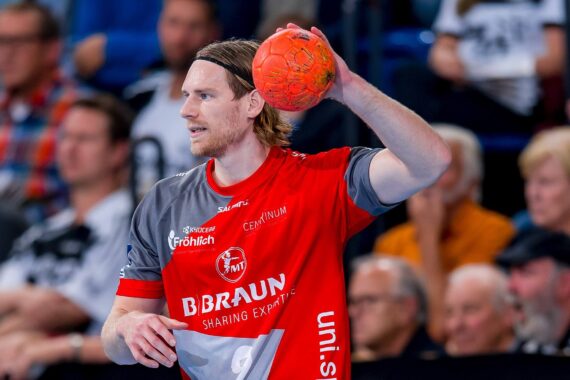 Ein männlicher Handballspieler in einem rot-grauen Trikot bereitet sich darauf vor, einen orangefarbenen Ball während eines Spiels zu werfen, während im Hintergrund Zuschauer sitzen.