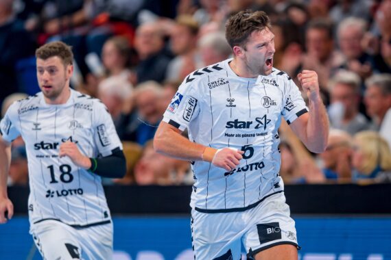 Zwei männliche Handballspieler in weißen Trikots feiern während eines Spiels, wobei der vordere Spieler die Faust ballt und vor Begeisterung schreit. Im Hintergrund ist eine unscharfe Zuschauermenge zu sehen.