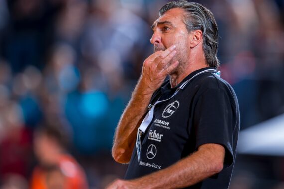 Ein Mann in einem schwarzen Polohemd mit Sponsorenlogos steht nachdenklich da und berührt mit der Hand sein Gesicht. Der Hintergrund ist unscharf und zeigt ein Publikum oder eine Menschenmenge in einer Außenumgebung.