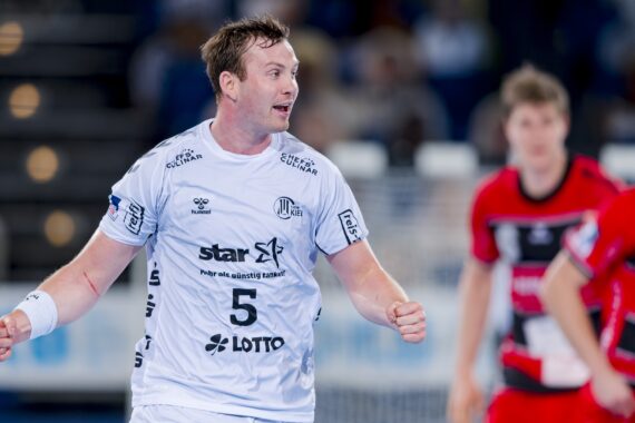 Ein männlicher Handballspieler in einem weißen Trikot mit der Nummer 5 steht auf dem Spielfeld und blickt mit leicht erhobenen Armen zur Seite. Zwei Spieler in roten Trikots sind im unscharfen Hintergrund zu sehen.