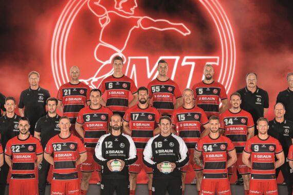 Eine Männerhandballmannschaft in roten und schwarzen Trikots posiert für ein Gruppenfoto mit Trainern und Betreuern vor einem roten Hintergrund mit einem großen Mannschaftslogo, das einen Handballspieler in Aktion zeigt.