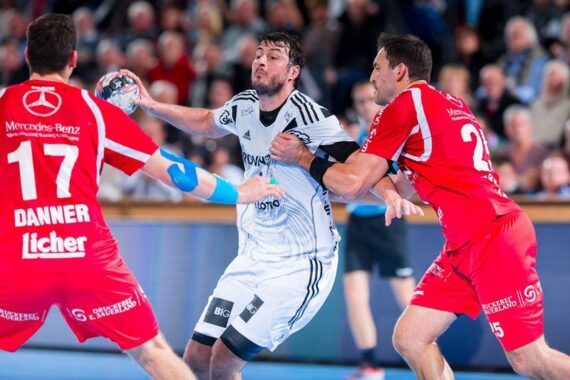 Ein männlicher Handballspieler in einem weißen Trikot versucht, während eines intensiven Spiels zwei Verteidiger in roten Trikots zu überwinden, während im Hintergrund eine unscharfe Zuschauermenge zusieht.