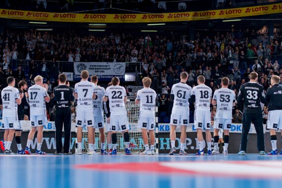 Eine Herren-Handballmannschaft steht in einer Reihe auf einem Hallenplatz vor einer jubelnden Menge, mit dem Rücken zur Kamera und in passenden weißen Trikots mit schwarzen Nummern und Sponsorenlogos.