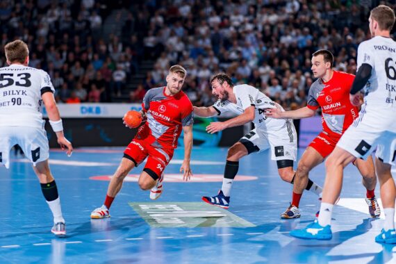 Ein Handballspiel der Männer in Aktion; ein Spieler in einem roten Trikot läuft mit dem Ball, während zwei Spieler in Weiß versuchen, ihn zu blockieren, mit weiteren Spielern und einer jubelnden Menge im Hintergrund.