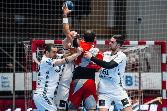 Ein Handballspieler in Rot springt zum Schuss, während drei Verteidiger in weißen Trikots ihn in der Nähe des Tors blockieren und alle ihre Arme nach oben strecken - ein intensiver Moment.