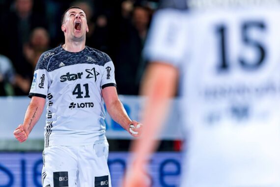Ein männlicher Handballspieler, der ein weißes Trikot mit der Nummer 41 trägt, schreit leidenschaftlich und feiert während eines Spiels. Ein anderer Spieler mit der Nummer 15 ist im Vordergrund zu sehen, leicht unscharf.