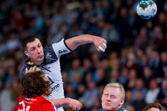Ein Handballspieler in einem grau-weißen Trikot springt und bereitet sich auf den Wurf vor, während zwei gegnerische Spieler in Rot und Grau versuchen, ihn während eines Spiels zu blockieren. Im Hintergrund ist eine verschwommene Menschenmenge zu sehen.