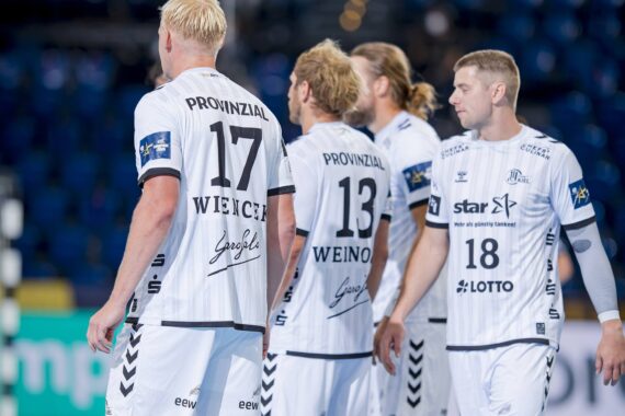 Vier männliche Handballspieler in weißen Trikots mit schwarzem Text und Sponsorenlogos stehen zusammen auf dem Spielfeld, mit dem Blick zur Seite und nach außen. Im Hintergrund sind unscharfe blaue Stadionsitze zu sehen.