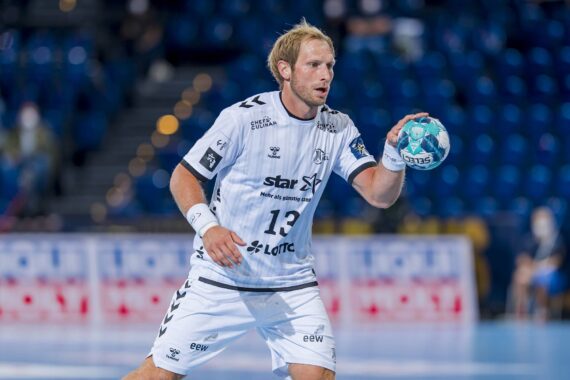 Ein männlicher Handballspieler in einem weißen Trikot mit der Nummer 13 bereitet sich darauf vor, einen blauen und grünen Ball während eines Spiels in einer Halle mit leeren blauen Sitzen im Hintergrund zu werfen.