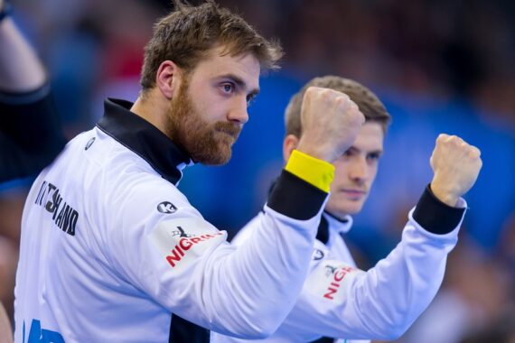 Zwei männliche Sportler in weißen Jacken heben ihre Fäuste in einer Geste der Entschlossenheit oder des Jubels und schauen konzentriert. Der Hintergrund ist unscharf, was darauf hindeutet, dass sie sich bei einer Sportveranstaltung oder einem Wettkampf befinden.