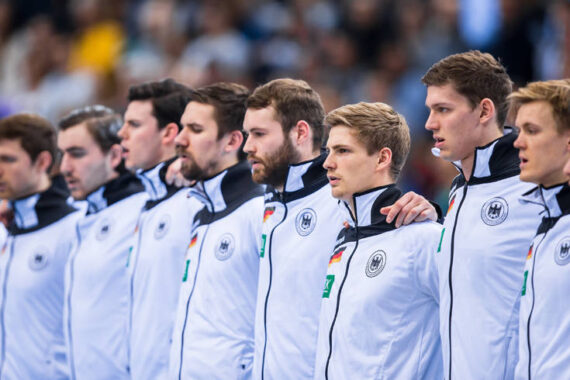 Eine Reihe männlicher Athleten in weißen deutschen Mannschaftsjacken stehen Seite an Seite und legen sich gegenseitig die Arme um die Schultern. Sie wirken konzentriert vor einer Sportveranstaltung. Im Hintergrund ist unscharf eine Menschenmenge zu sehen.