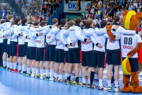 Eine Handballmannschaft steht Seite an Seite mit den Armen um die Schultern des anderen, von der Kamera abgewandt. Eine Person in einem Bärenmaskottchenkostüm steht am Ende der Reihe. Die Zuschauer füllen die Tribünen im Hintergrund.