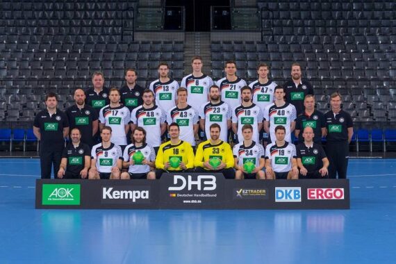 Eine Herren-Handballmannschaft in schwarz-weißen Trikots stellt sich für ein Gruppenfoto in einer Halle auf. Trainer und Betreuer stehen auf beiden Seiten, und drei Spieler in gelben Trikots sitzen vorne und halten grüne Handbälle.