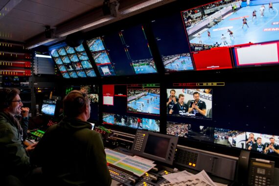 Zwei Personen sitzen an einer Schalttafel in einem Fernsehstudio und überwachen mehrere Bildschirme, auf denen ein Sportereignis und die Kommentatoren live gezeigt werden. Der Raum ist voll mit Monitoren, Schalttafeln und technischen Geräten.