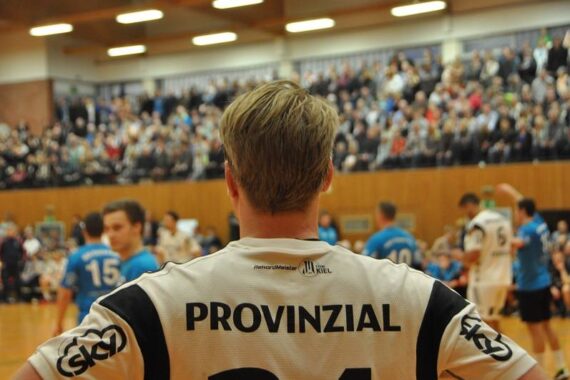 Ein Handballspieler, der ein weißes Trikot mit der Aufschrift PROVINZIAL trägt, steht während eines Spiels mit den Händen in den Hüften vor einer überfüllten Sporthalle und anderen Spielern in blauen Trikots.