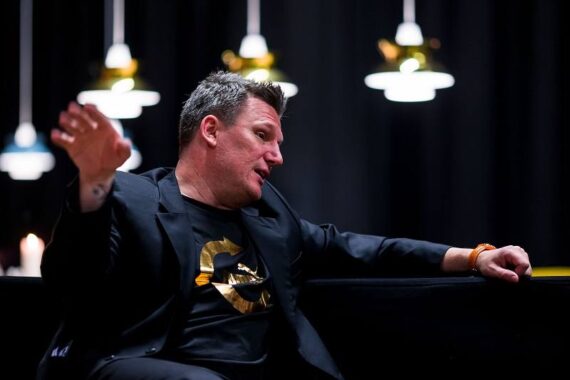 Ein Mann in schwarzem Anzug und T-Shirt gestikuliert mit dem Arm, während er in einem Innenraum sitzt, mit modernen Hängelampen und einem dunklen Hintergrund hinter ihm.