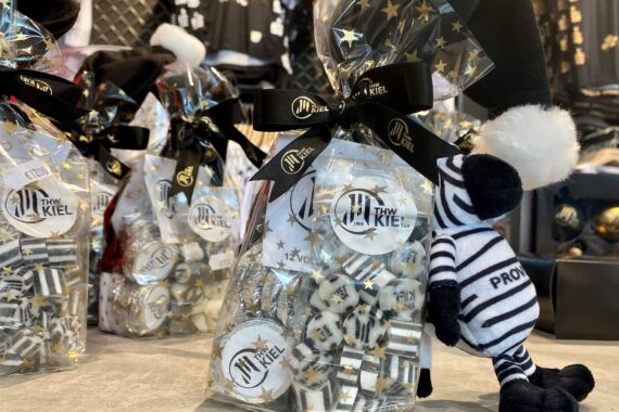 Auf einem Tresen stehen Geschenktüten mit schwarzen Bändern und goldenen Sternen, die mit THW Kiel beschriftet sind. Neben den Tüten steht ein kleines Plüsch-Zebra im gestreiften Outfit und mit weißer Bommelmütze.