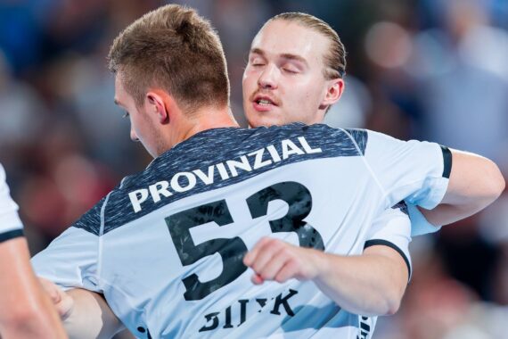 Zwei männliche Handballspieler in weißen und schwarzen Trikots umarmen sich während eines Spiels; das Trikot des einen Spielers trägt die Nummer 53 und PROVINZIAL auf dem Rücken. Der Hintergrund ist unscharf und lässt eine Sportarena vermuten.