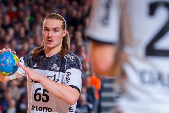 Ein männlicher Handballspieler mit langen Haaren bereitet sich darauf vor, einen bunten Ball während eines Spiels zu passen, mit einem verschwommenen Mitspieler im Vordergrund und einer Menschenmenge im Hintergrund.
