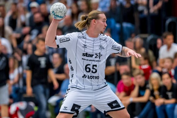 Ein männlicher Handballspieler in einem weißen Trikot bereitet sich darauf vor, den Ball während eines Spiels zu werfen, während im Hintergrund eine Menge Zuschauer zusieht.