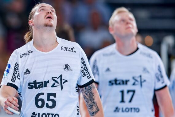 Zwei männliche Handballspieler in weißen Trikots mit den Nummern 65 und 17 blicken während eines Spiels mit konzentrierter Miene nach oben. Der Hintergrund ist unscharf und hebt die Sportler im Vordergrund hervor.