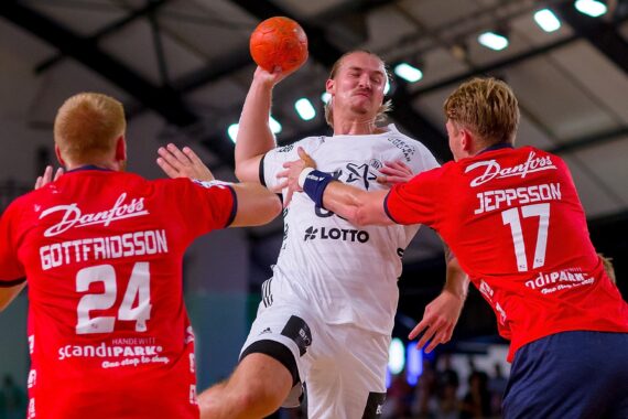 Ein Handballspieler in einem weißen Trikot springt, um den Ball zu schießen, während zwei Verteidiger in roten Trikots versuchen, ihn während eines intensiven Spiels in einer Halle zu blockieren.