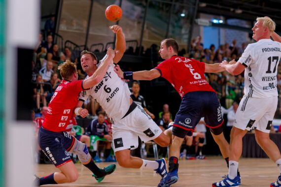 Vier männliche Handballspieler, zwei in Weiß und zwei in Rot, sind auf einem Hallenplatz in heftiger Aktion. Ein weiß gekleideter Spieler springt und versucht einen Schuss, während er von zwei rot gekleideten Verteidigern geblockt wird.