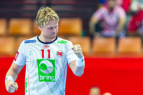 Ein männlicher Handballspieler in einem weißen Trikot mit der Nummer 11 und der norwegischen Flagge ballt auf dem Spielfeld jubelnd die Faust. Im Hintergrund sind unscharfe Zuschauer in einer Sportarena zu sehen.