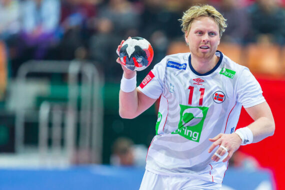 Ein männlicher Handballspieler in einem weißen norwegischen Trikot mit der Nummer 11 hält den Ball und bereitet sich auf den Wurf während eines Spiels vor. Der Hintergrund ist unscharf und zeigt Zuschauer und das Spielfeld.