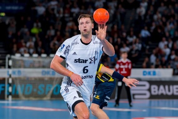 Ein männlicher Handballspieler in einem weißen Trikot mit der Nummer 6 bereitet sich darauf vor, einen orangefarbenen Ball während eines Spiels zu fangen. Ein anderer Spieler in einem dunklen Trikot und ein Torwart sind im Hintergrund auf einem blauen Spielfeld zu sehen.