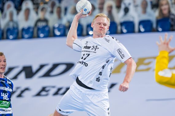 Ein Handballspieler in einem weißen Trikot springt, um den Ball in Richtung Tor zu werfen, während ein Spieler in Blau und ein Torwart in Gelb sich auf die Verteidigung vorbereiten. Im unscharfen Hintergrund sind Zuschauer zu sehen.