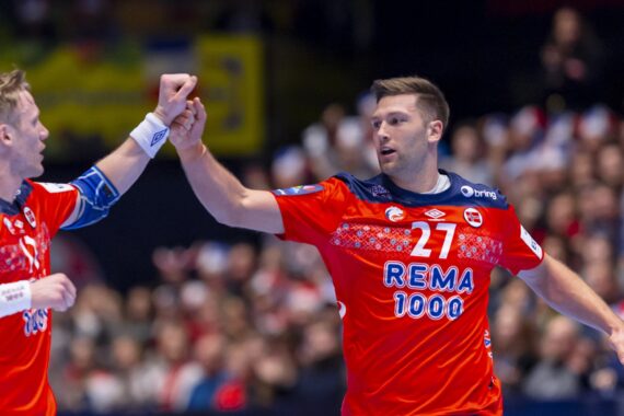 Zwei männliche Handballspieler in roten norwegischen Trikots feiern während eines Spiels mit einem Faustschlag. Einer trägt die Nummer 27. Im Hintergrund ist eine unscharfe Menschenmenge zu sehen.