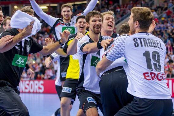 Eine Gruppe männlicher Handballspieler in schwarz-weißen Trikots feiert ausgelassen auf dem Spielfeld, umarmt sich und lächelt nach einem Sieg. Der Spieler im Vordergrund hat die Strobel 19 auf dem Rücken. Im Hintergrund ist die Zuschauermenge zu sehen.