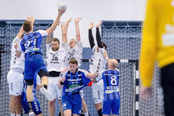 Ein Handballspieler in einem blauen Trikot springt zum Schuss, während mehrere Verteidiger in weißen Trikots in der Nähe des Tors aufspringen, um zu blocken, während andere Spieler und ein Torwart in der Szene zu sehen sind.