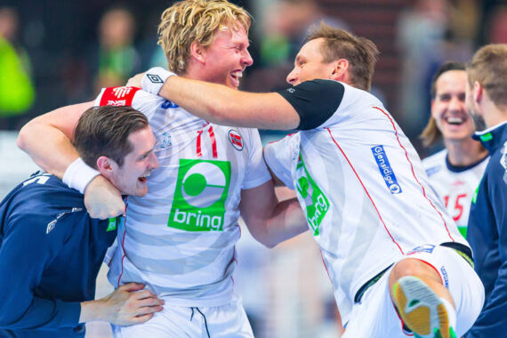Drei männliche Handballspieler in weißen Trikots feiern ausgelassen und umarmen sich auf dem Spielfeld. Sie lächeln und scheinen aufgeregt zu sein, wobei ein Spieler mitten im Sprung ist, während andere im Hintergrund energisch jubeln.