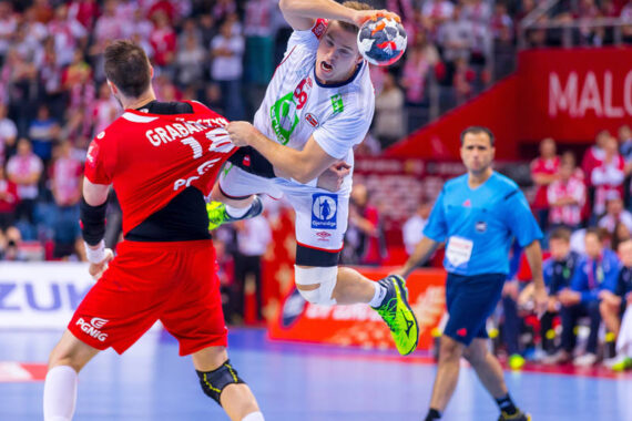 Ein Handballspieler in einem weißen Trikot springt, um den Ball zu schießen, während er von einem Spieler in einem roten Trikot verteidigt wird. Zuschauer und Offizielle sind im Hintergrund auf dem Hallenplatz zu sehen.