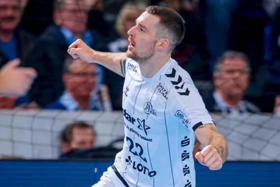 Ein männlicher Handballspieler in einem weißen Trikot mit der Nummer 22 jubelt während eines Spiels, während das Publikum im Hintergrund verschwommen zu sehen ist.