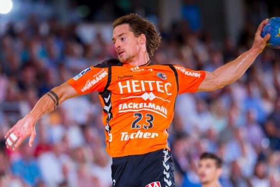 Ein männlicher Handballspieler, der ein orangefarbenes Trikot mit der Aufschrift HEITEC und der Nummer 23 trägt, springt mit erhobenem Arm in die Luft und bereitet sich darauf vor, einen Ball zu werfen. Im Hintergrund ist eine unscharfe Menschenmenge zu sehen.