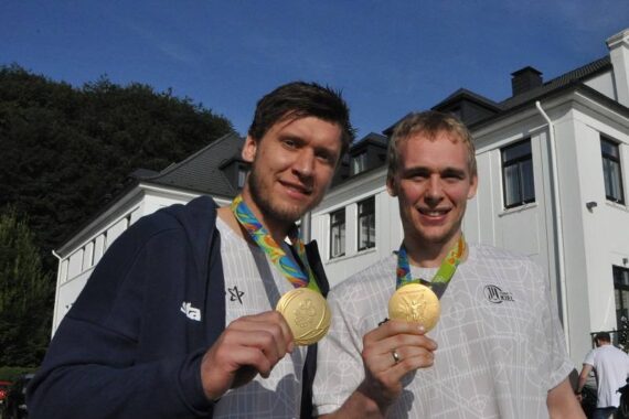 Zwei Sportler in weißen Hemden lächeln und halten Goldmedaillen hoch, während sie an einem sonnigen Tag vor einem großen weißen Gebäude stehen.