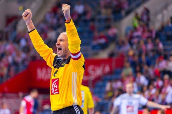 Ein Handballtorwart in einem gelben Trikot jubelt mit erhobenen Armen und wirkt während eines Spiels auf dem Spielfeld freudig und triumphierend, während Spieler und Zuschauer im Hintergrund verschwommen zu sehen sind.