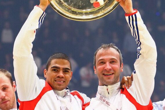 Zwei Athleten in weiß-roten Trainingsanzügen feiern mit Medaillen und halten eine große goldene Trophäenplatte hoch.