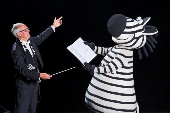 Ein Mann im Smoking dirigiert eine Person in einem großen Zebrakostüm, die Notenblätter in der Hand hält, beide stehen vor einem schwarzen Hintergrund.